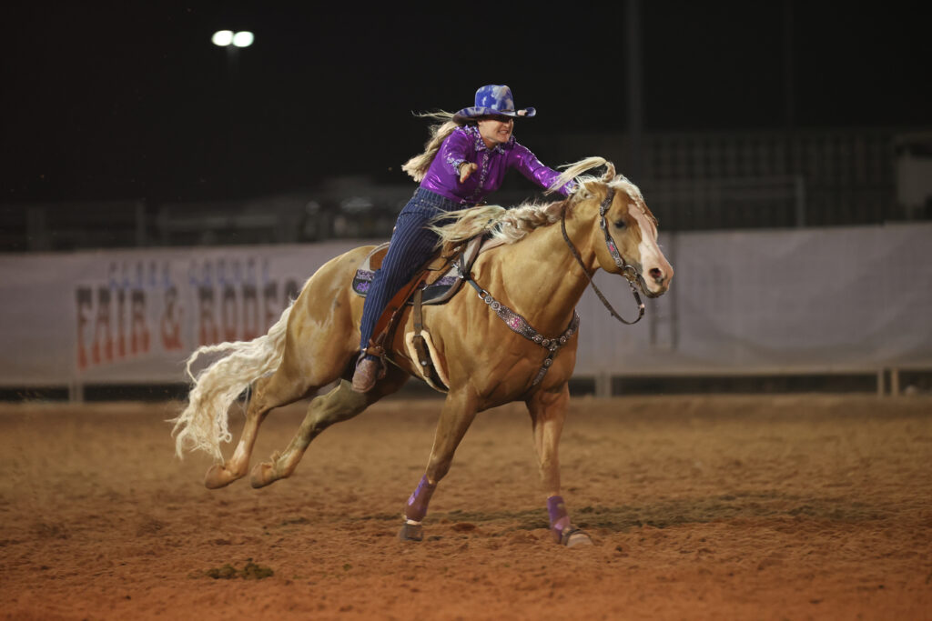 Barrel racer in a purple shirt and blue hat gallops her palomino horse across the arena dirt under the lights
