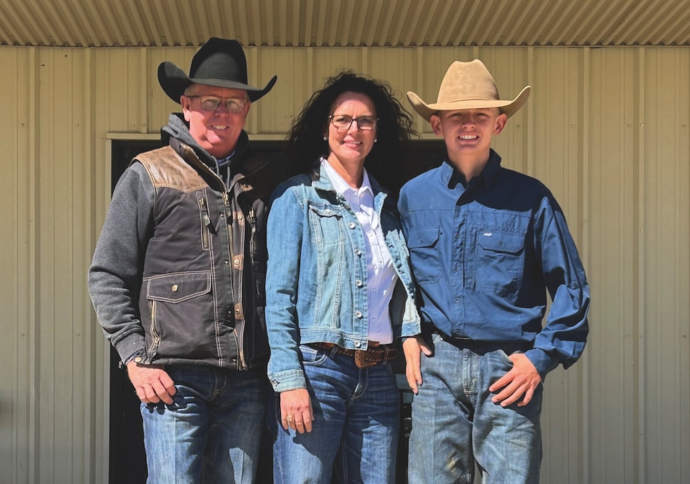 Three family members in cowboy hats, denim and work jackets stand smiling together in front of a metal barn wall
