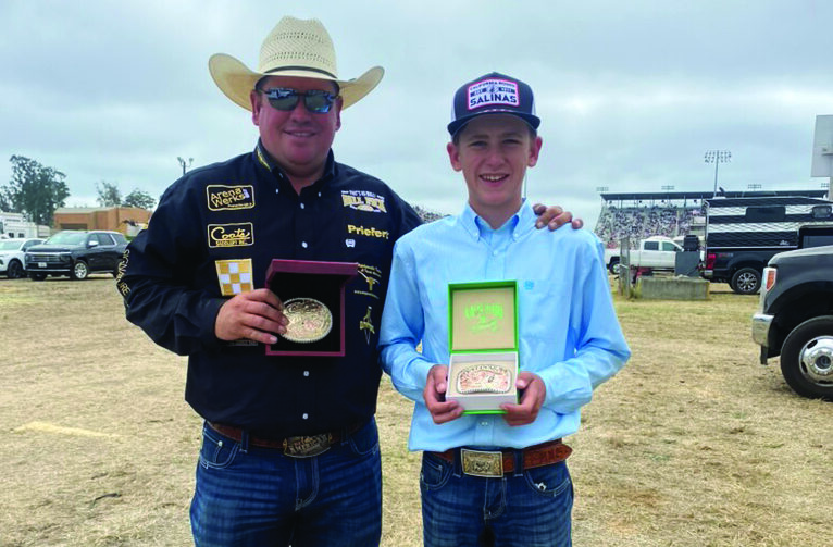 A man in a straw cowboy hat and black sponsor shirt stands with a young roper in a Salinas cap, both holding championship buckles at a rodeo grounds