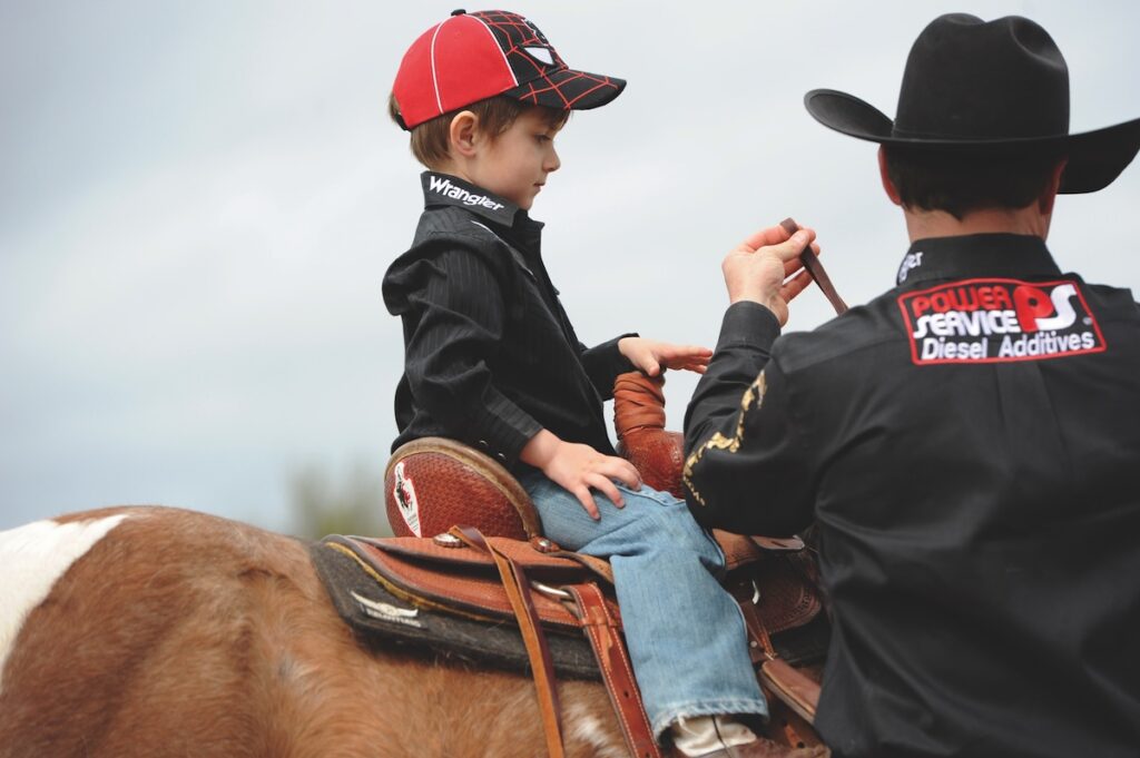 Young boy in a red ball cap and black Wrangler shirt sits in a western saddle as a cowboy hands him the reins