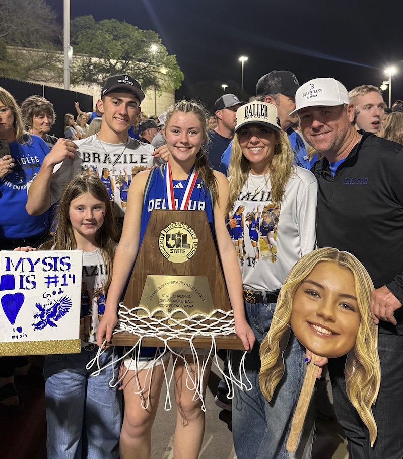 Family gathers around a girl in a Decatur jersey holding a UIL state champion basketball trophy with a cut-down net