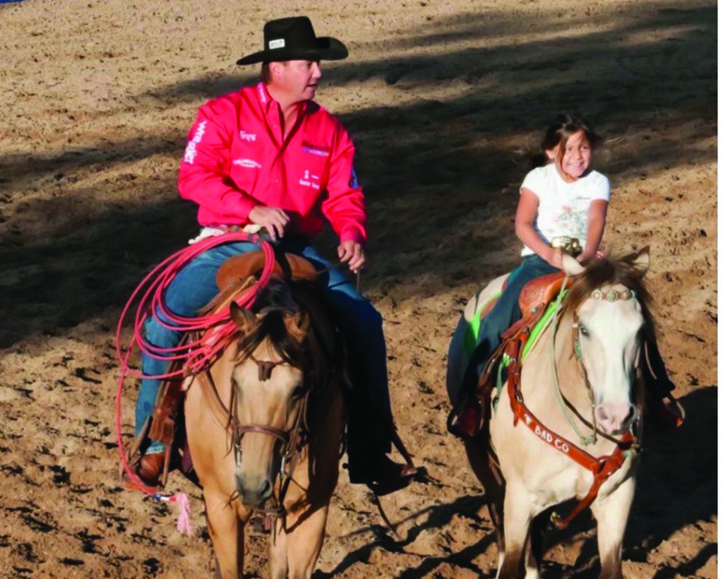 Cowboy in a pink shirt and black hat rides alongside a smiling young girl on a buckskin horse in a dirt arena