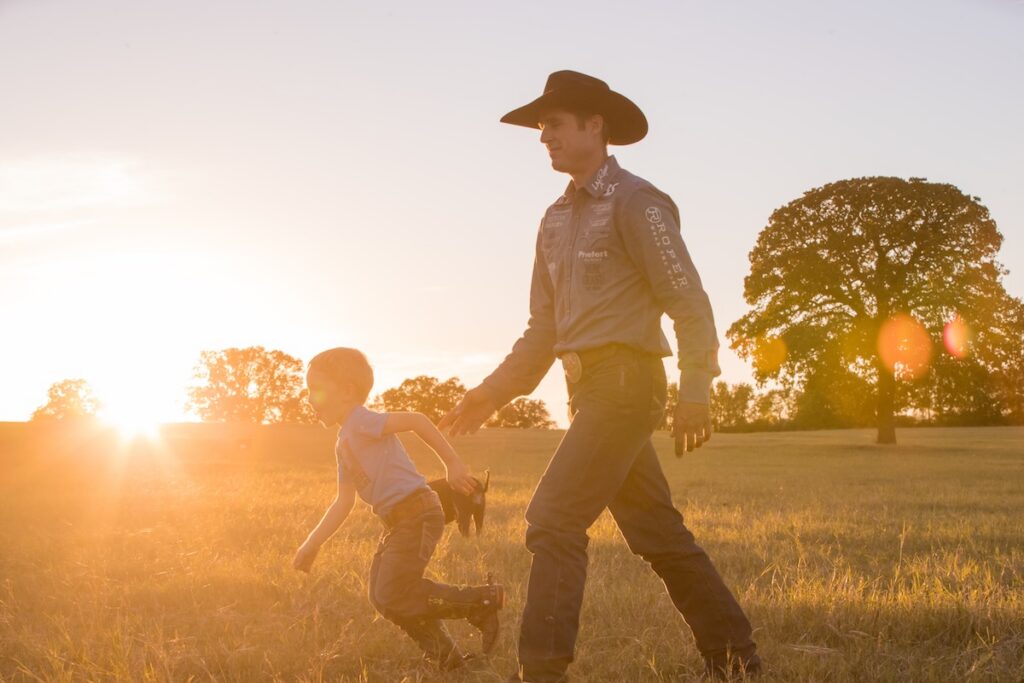 Cowboy in a black hat and Roper shirt walks across a sunlit pasture with a young boy running alongside him