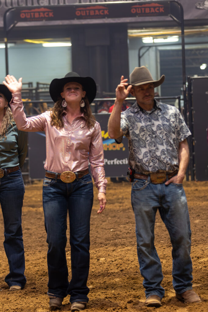 Contestants in cowboy hats wave to the crowd on the dirt arena floor during rodeo introductions