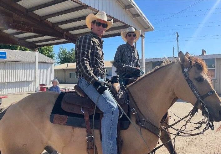 Two riders in cowboy hats and sunglasses sit horseback on buckskin horses under a covered arena alley