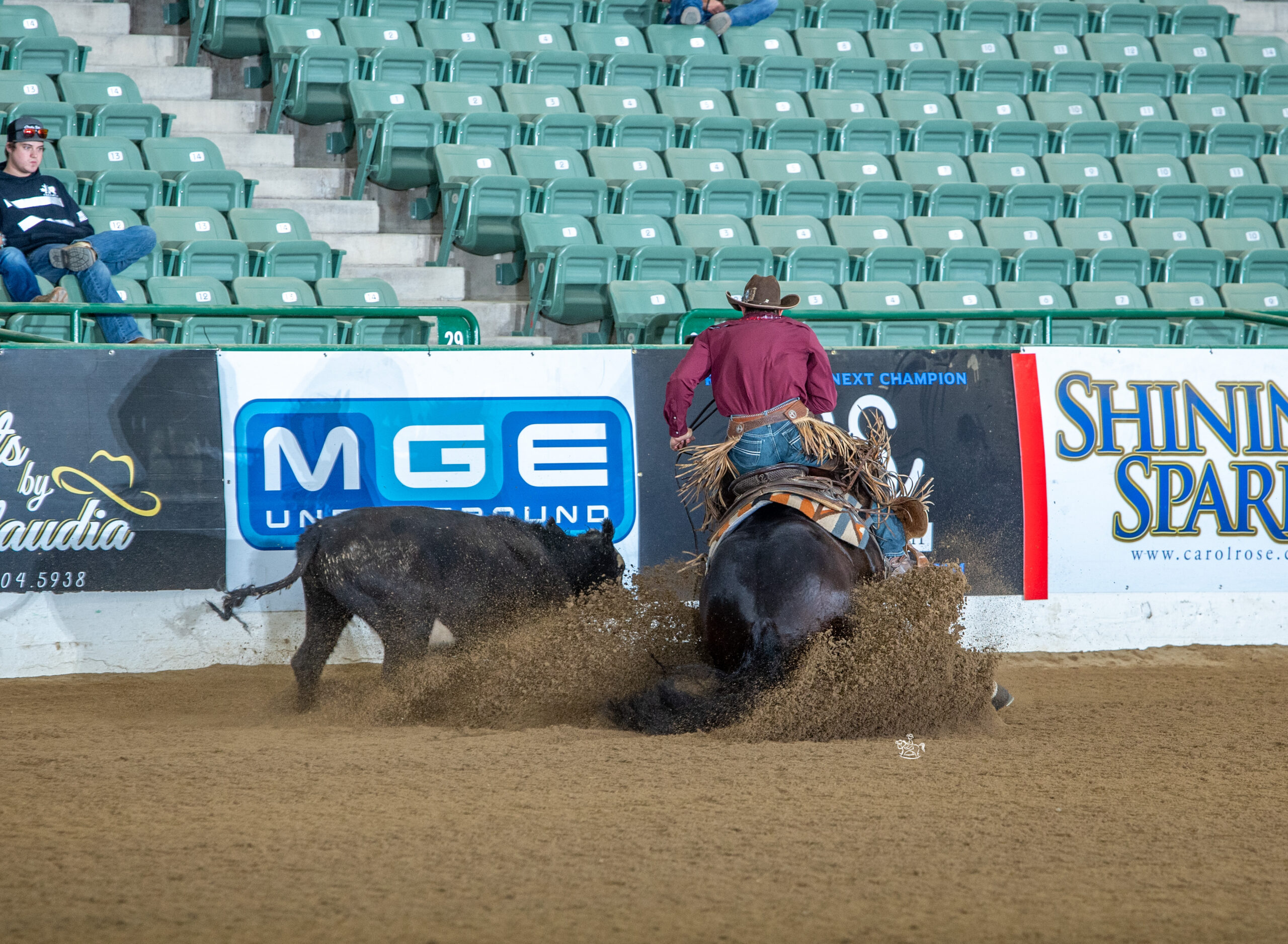Cowboy in maroon shirt slides a black horse to a stop in the arena dirt, kicking up dust near the barrier boards