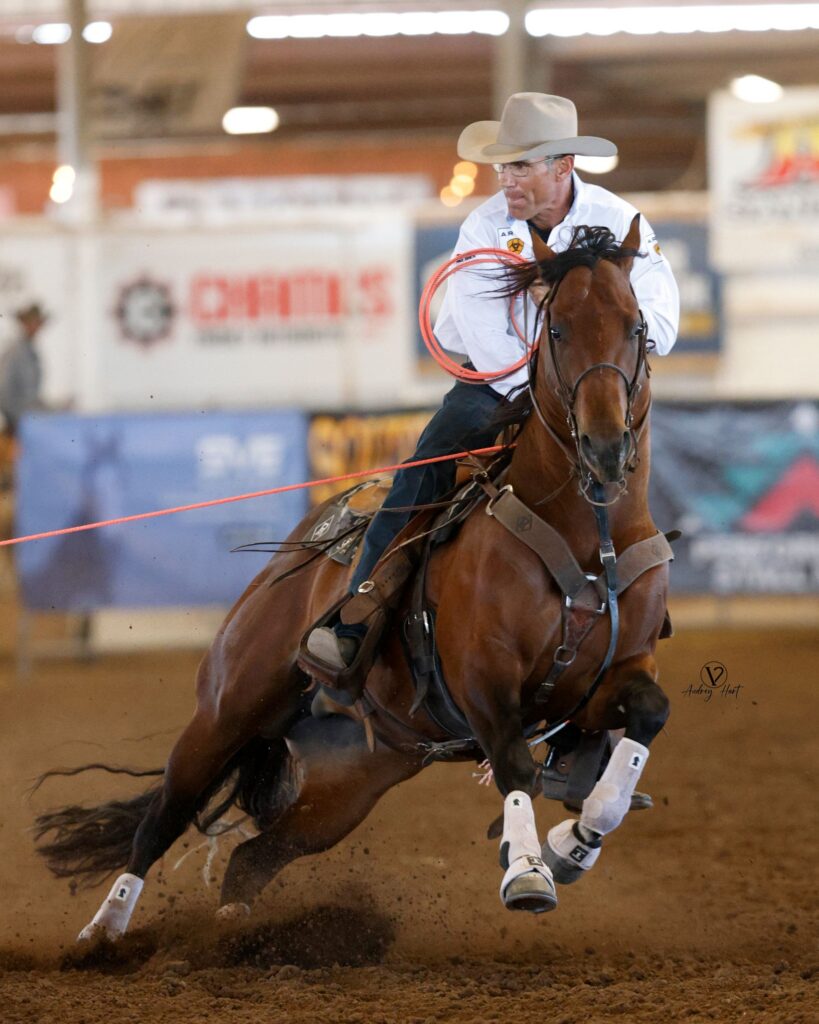 Roper on brown horse carrying coiled lasso gallops across indoor arena dirt during competition