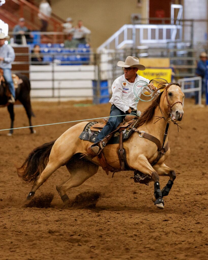 Roper on palomino horse galloping across arena dirt with lasso, wearing white shirt and cowboy hat during rodeo competition