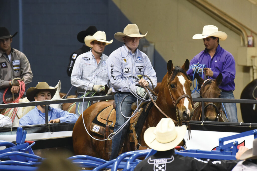 Team roper in a cowboy hat holds his coiled rope while sitting horseback behind the arena as other cowboys wait nearby
