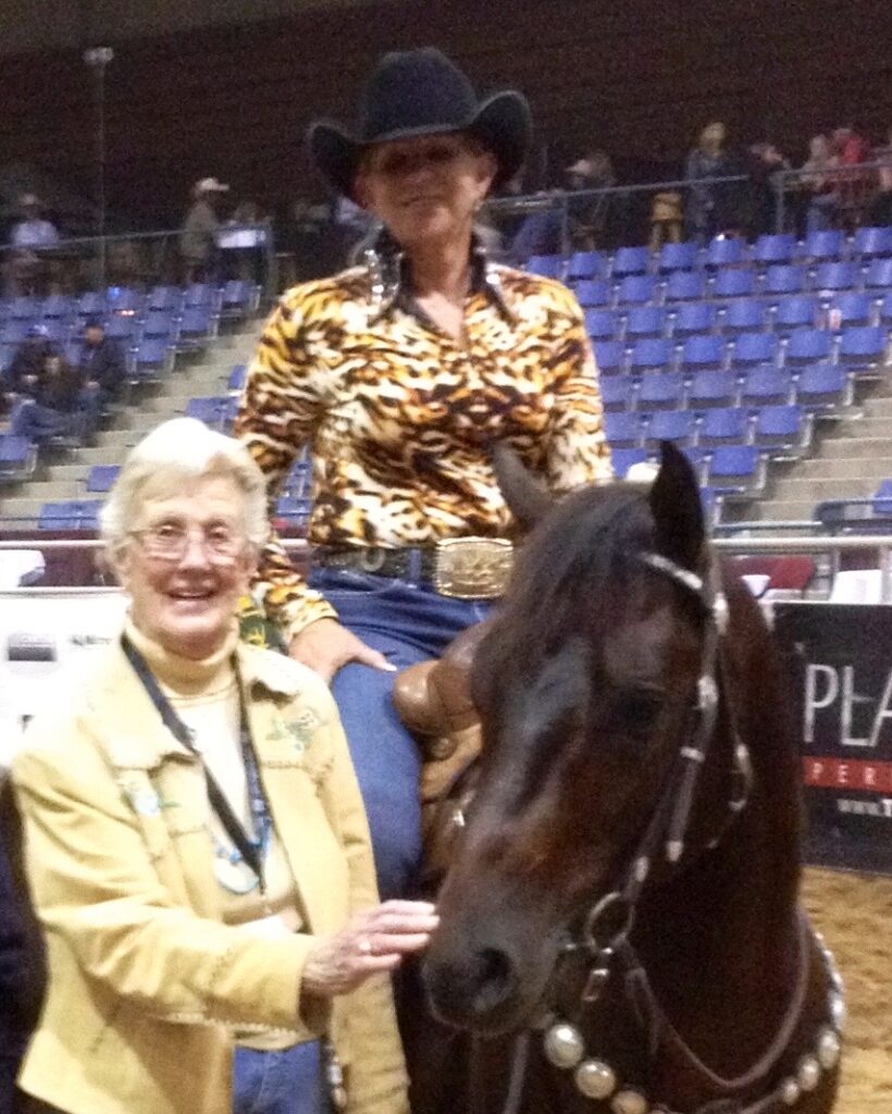 Smiling rider in a black cowboy hat and animal-print show shirt sits astride a dark bay horse while an older woman stands beside them in an arena