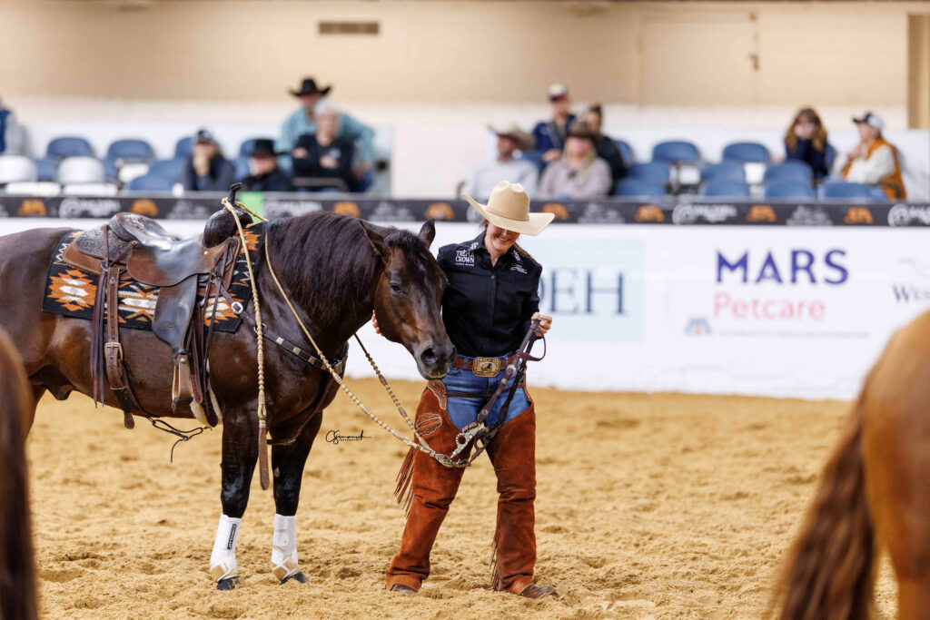 Exhibitor in a felt cowboy hat and chaps smiles while leading her saddled bay horse in an indoor arena
