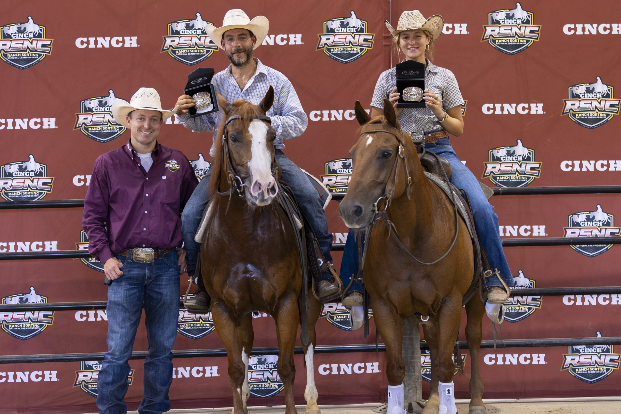 Three rodeo competitors stand on a podium with their horses wearing cowboy hats at a Cinch-sponsored event