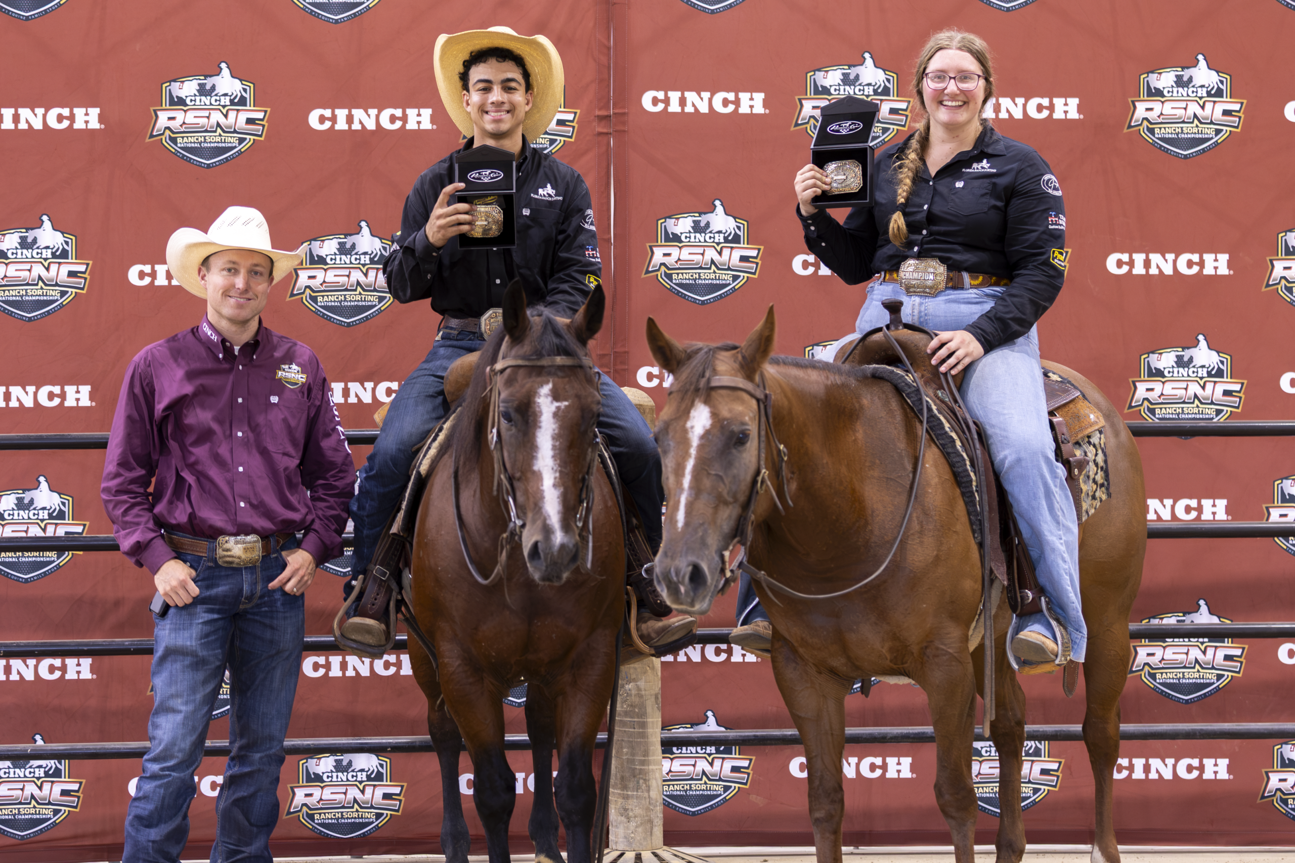 Two mounted riders in Western attire pose with handlers at a Cinch RSNC event backdrop during an awards presentation