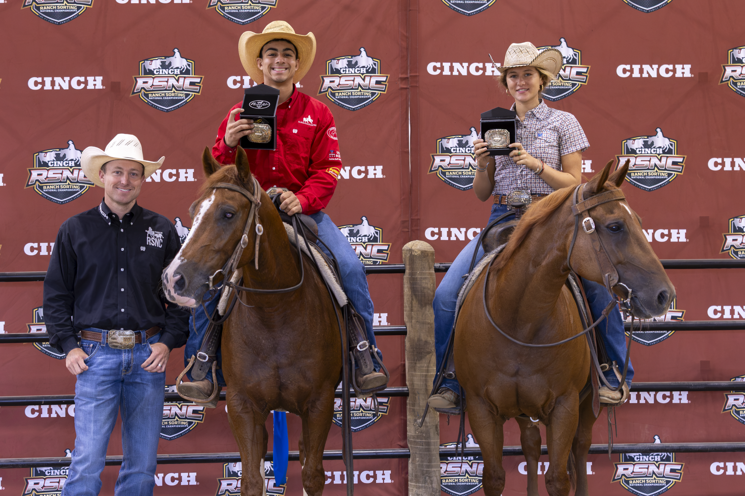 Rodeo competitors in western attire stand with two saddled horses at a Cinch-branded event backdrop during an awards presentation