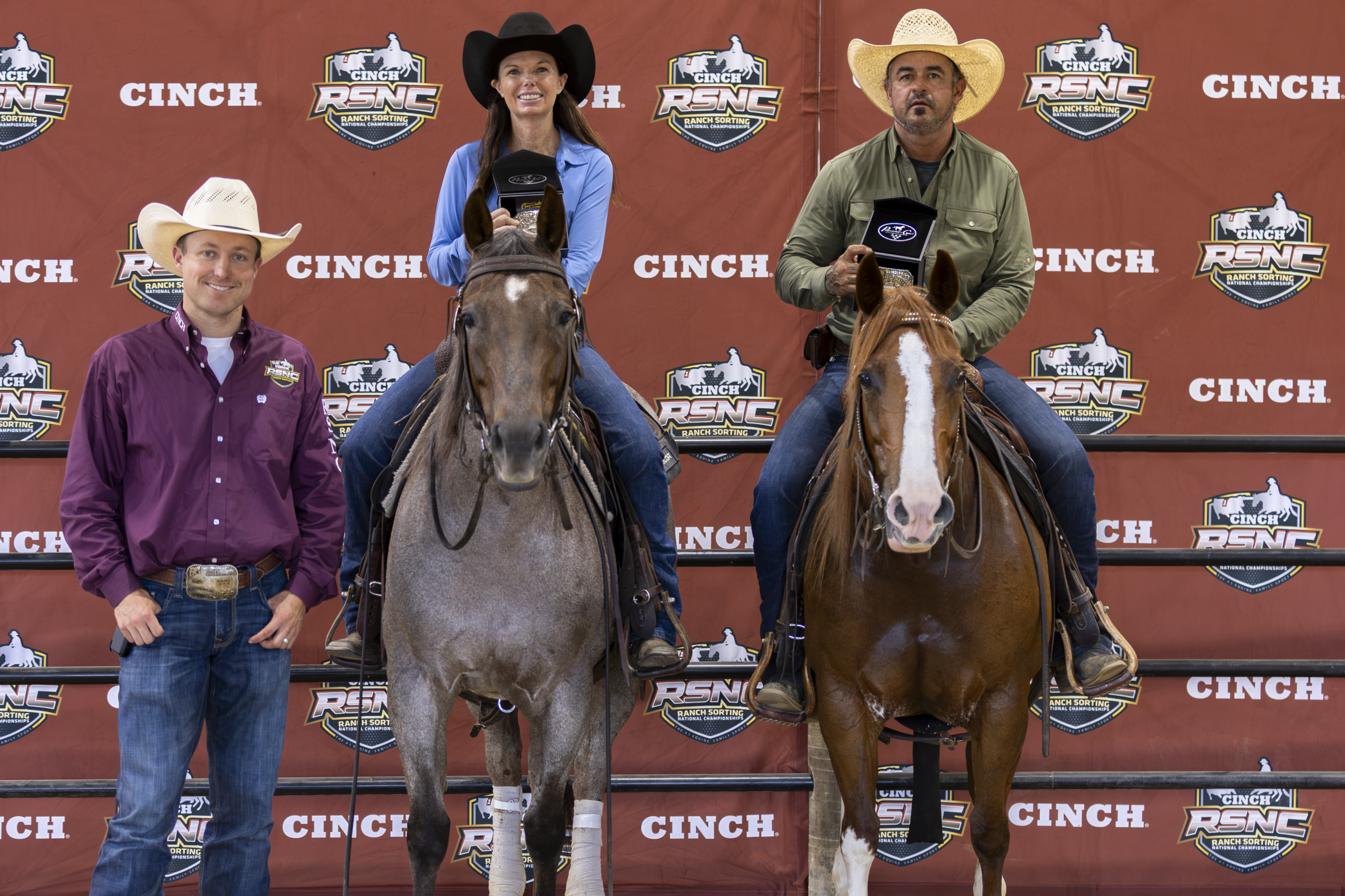 Three cowboys with horses stand before a Cinch-branded backdrop at a rodeo event