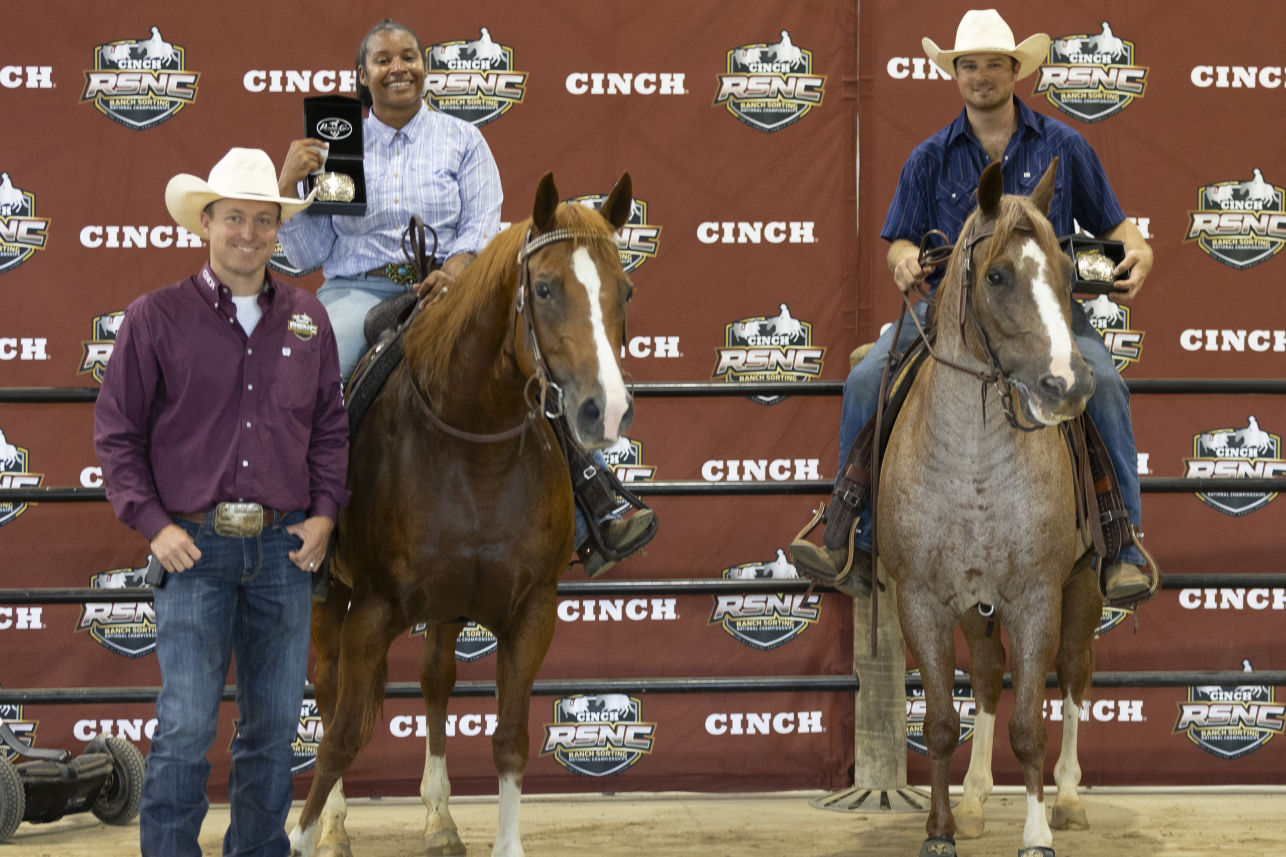 Four people in cowboy hats stand with two saddled horses at a Cinch-branded rodeo backdrop during an awards presentation