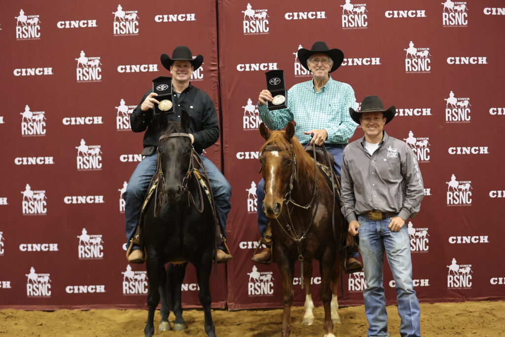 Two mounted riders hold up championship buckles beside a man standing on the ground, in front of a Cinch RSNC backdrop