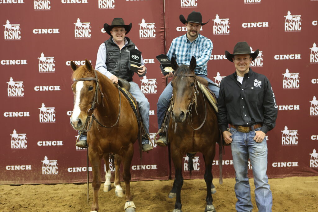 Two mounted cowboys hold award buckles and a plaque while a third man stands beside them in front of a Cinch RSNC backdrop