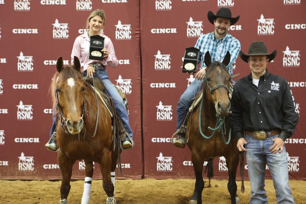 Two mounted riders hold up buckle presentation cases beside a standing man in front of a Cinch RSNC backdrop