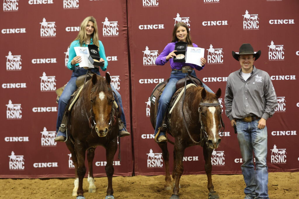 Two young riders on horseback hold award plaques and buckles beside a man in a cowboy hat at a Cinch RSNC backdrop