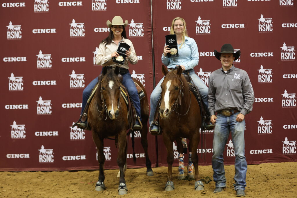 Two mounted riders hold buckle awards beside a man in a cowboy hat in front of a Cinch RSNC backdrop
