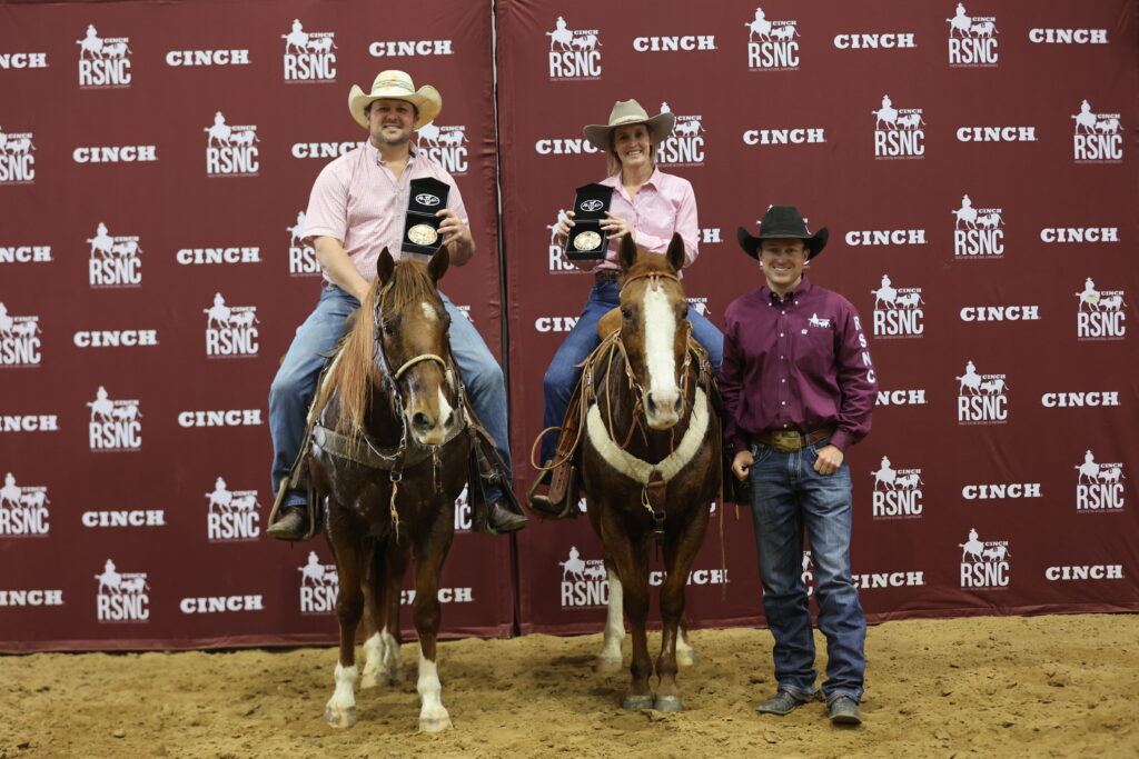 Two riders on horseback hold up trophy buckles beside a standing man in front of a Cinch RSNC backdrop