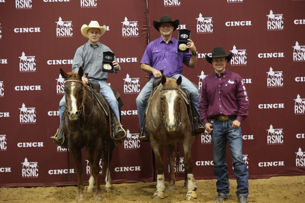Two mounted riders hold up belt buckles beside a man standing on the ground in front of a Cinch RSNC backdrop