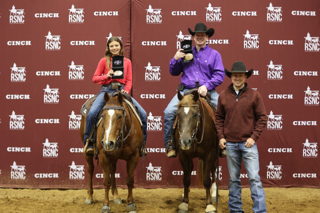 Two mounted riders hold up championship buckles beside a man standing at a Cinch RSNC backdrop
