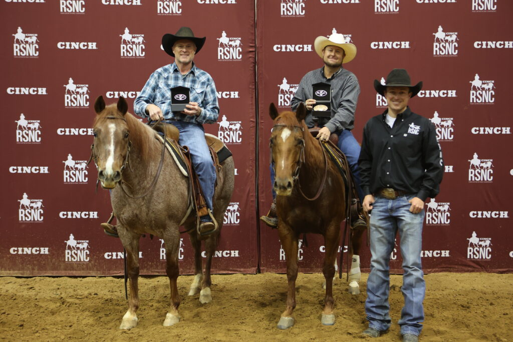 Two mounted cowboys hold buckle cases beside a standing man in front of a Cinch RSNC backdrop in an arena