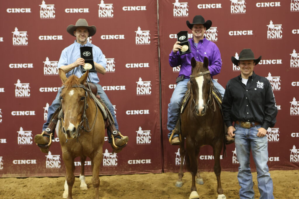 Two mounted cowboys hold up buckle cases beside a man standing on the ground in front of a Cinch RSNC backdrop
