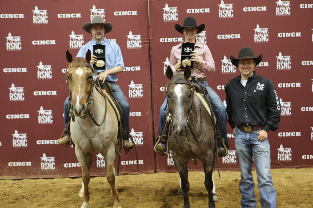 Two mounted riders in cowboy hats hold up champion buckles beside a man standing in front of a Cinch RSNC backdrop