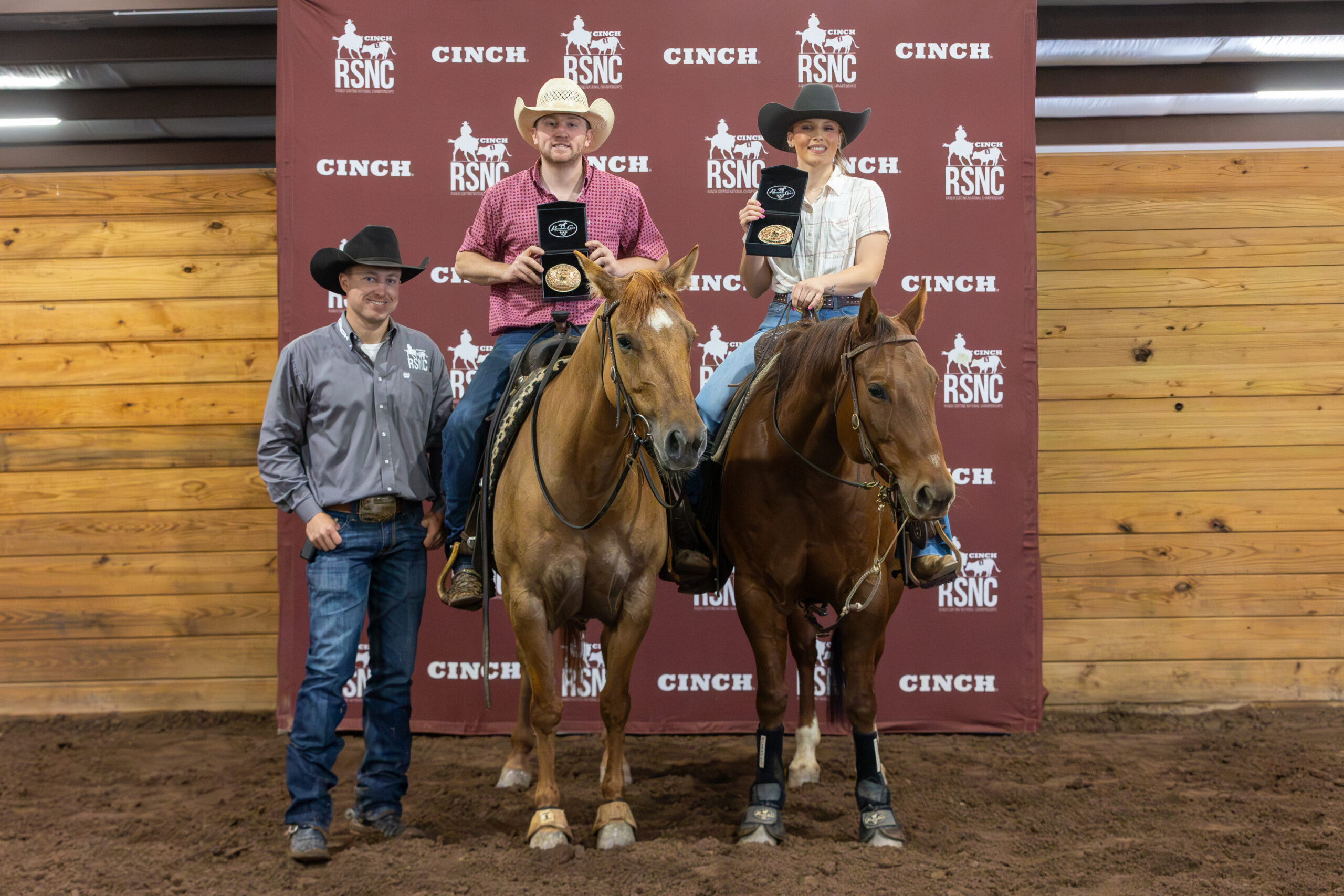 Two mounted riders hold up championship buckles beside a standing man in a cowboy hat before a Cinch RSNC backdrop