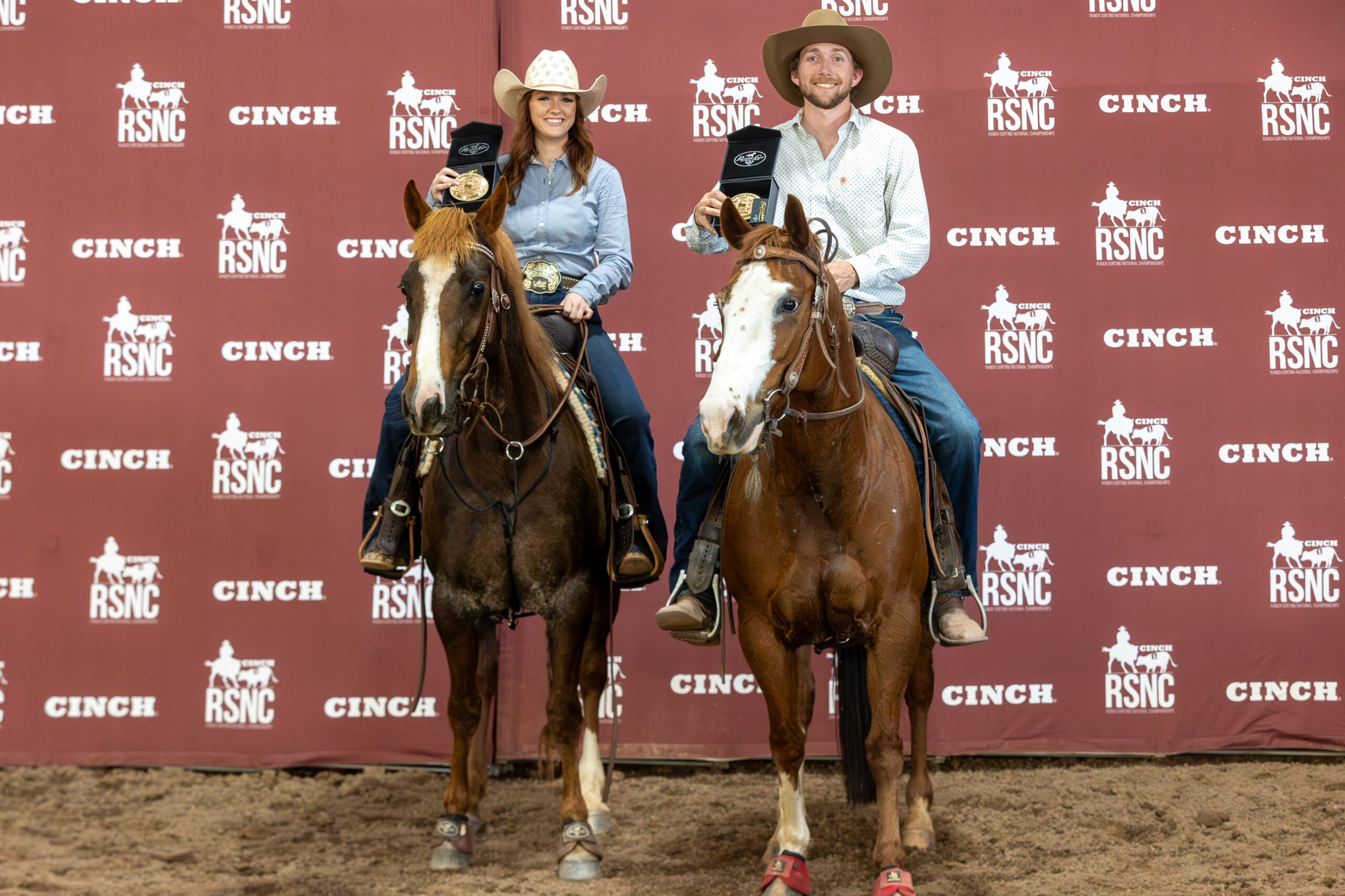 Two mounted riders in cowboy hats hold up championship buckles in presentation boxes before a Cinch RSNC backdrop