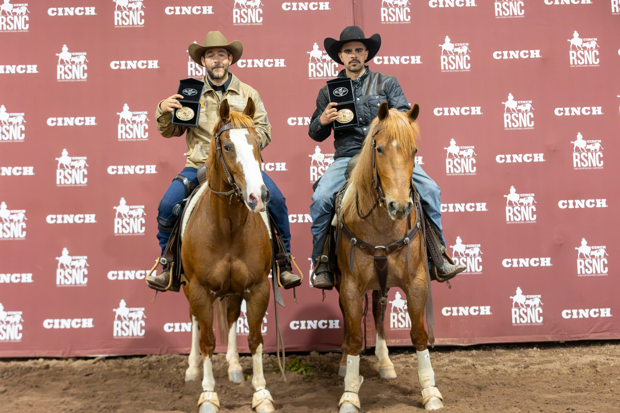 Two mounted cowboys hold up championship buckles in presentation boxes in front of a Cinch RSNC backdrop