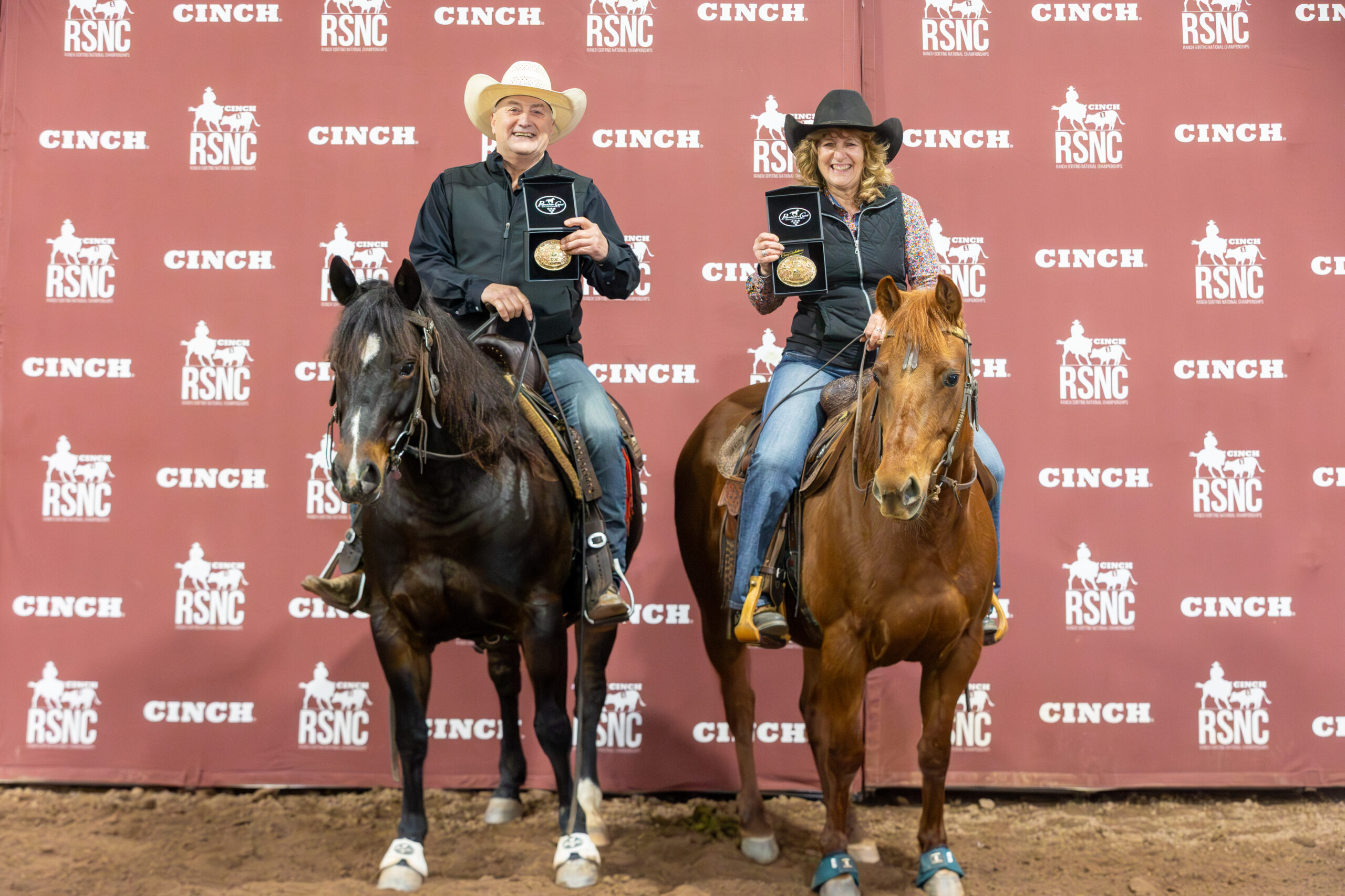 Two mounted riders smile while holding up gold championship buckles in front of a Cinch RSNC backdrop