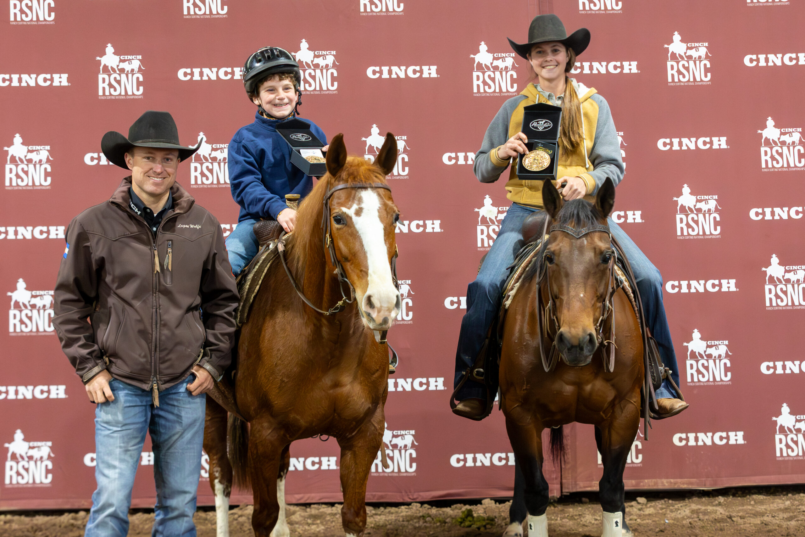 Young rider in a helmet and a woman in a cowboy hat hold up buckle boxes on horseback beside a standing cowboy at an RSNC backdrop
