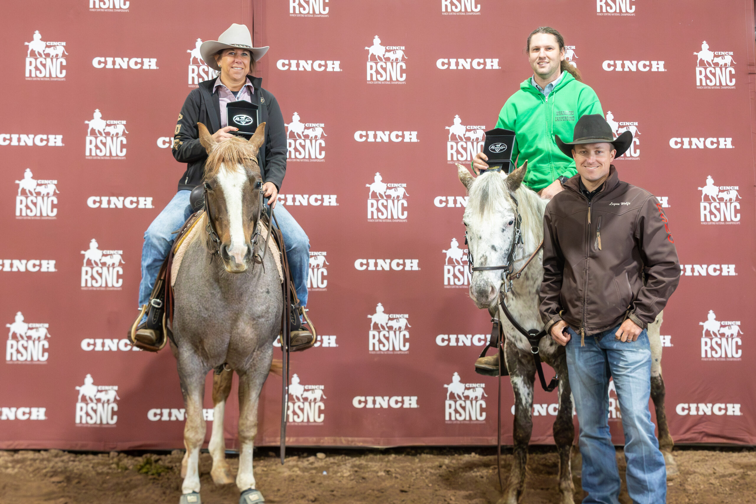 Two mounted riders hold buckle cases beside a man in a black cowboy hat in front of a Cinch RSNC backdrop