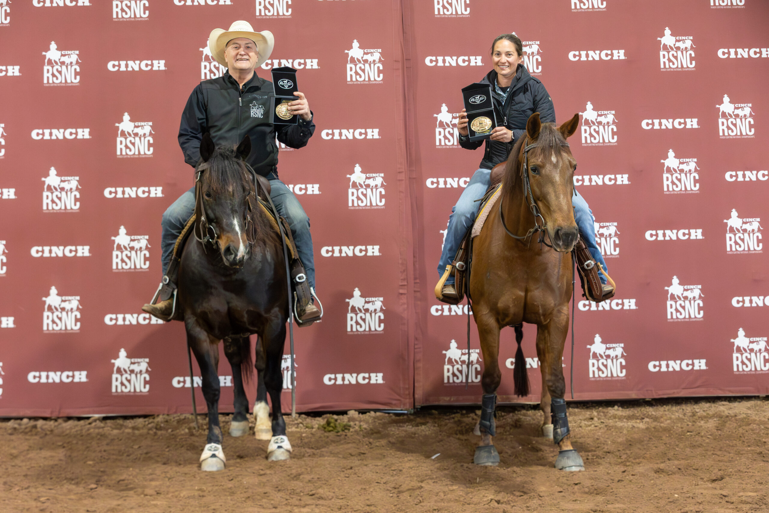 Two mounted riders hold up championship buckles in presentation boxes in front of a Cinch RSNC backdrop