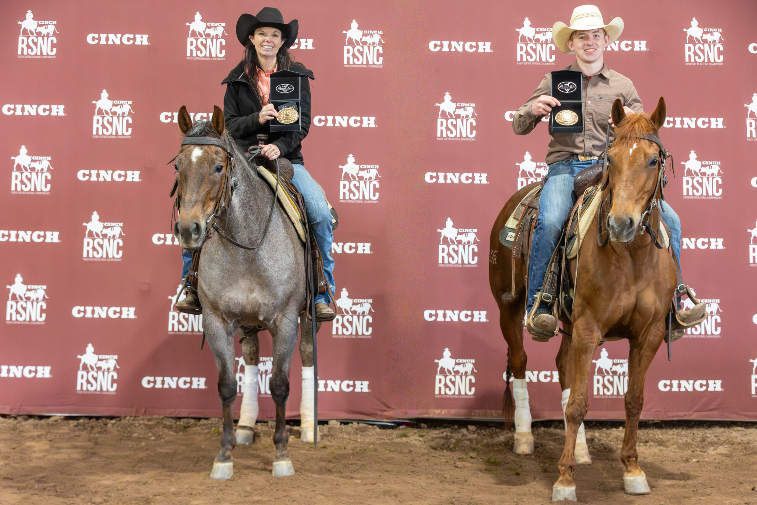 Two riders on horseback hold up championship buckles in presentation boxes in front of a Cinch RSNC backdrop