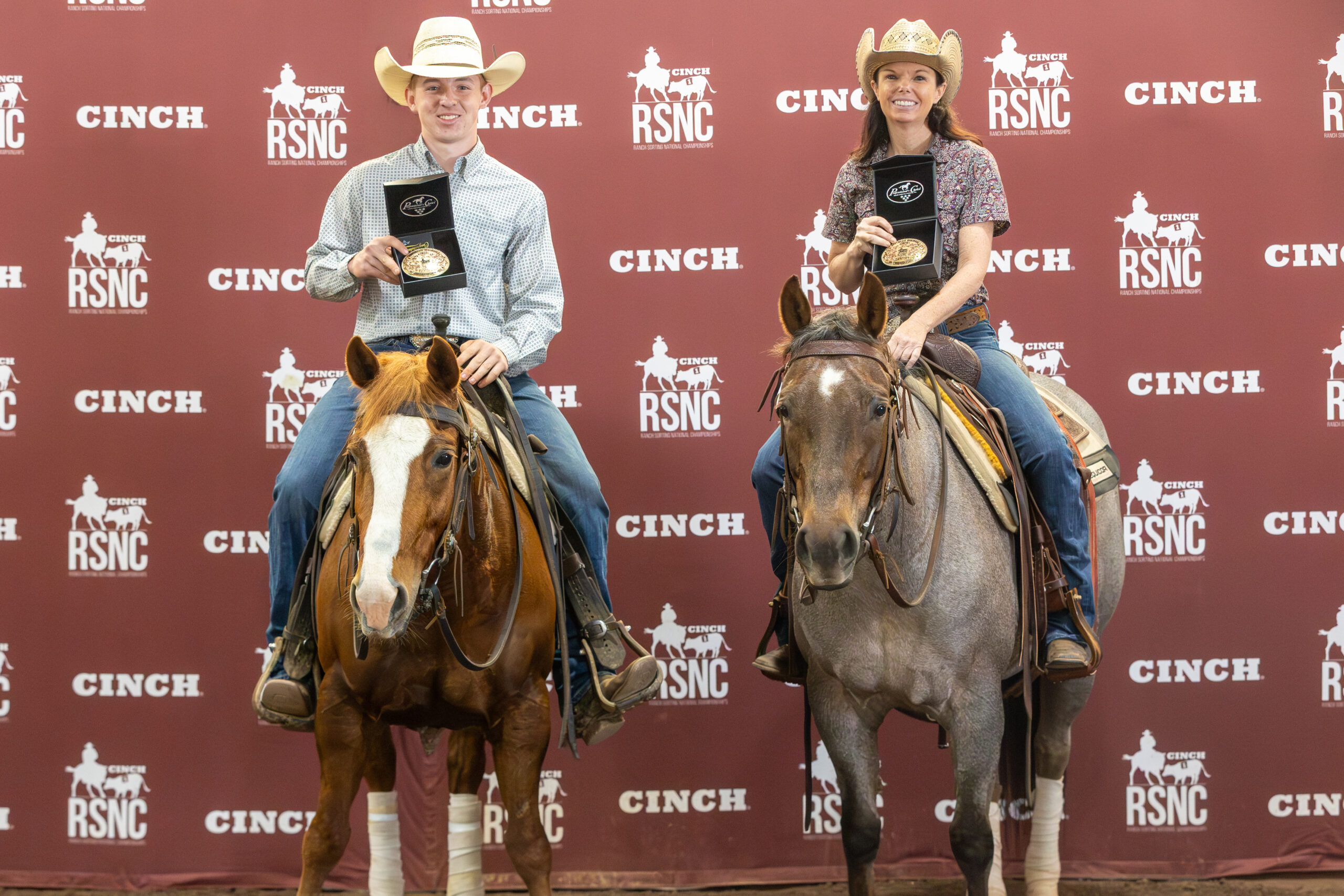 Two mounted riders hold up championship buckles in presentation boxes in front of a Cinch RSNC backdrop