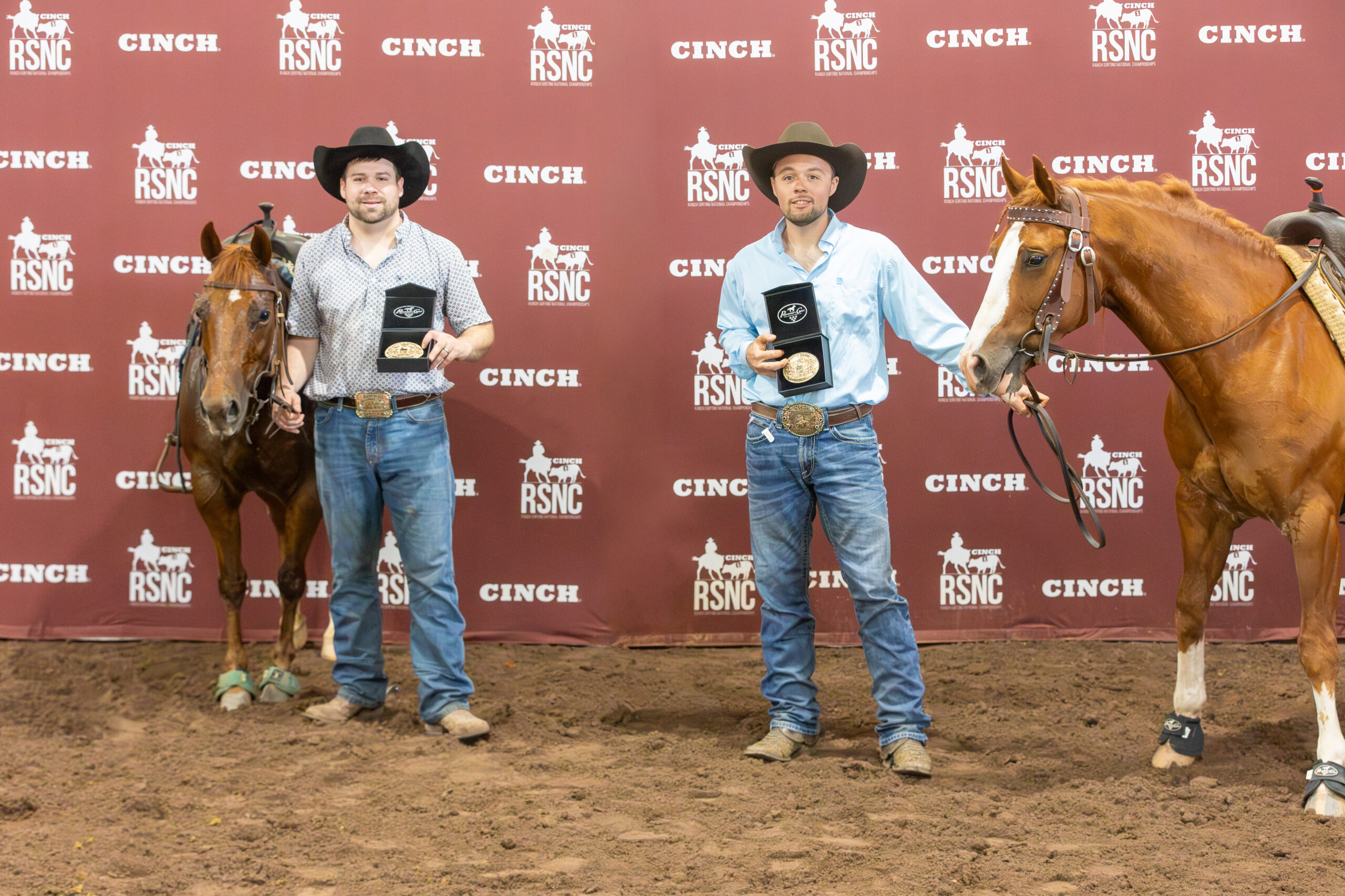 Two cowboys in black hats hold champion buckles in presentation boxes beside their sorrel horses at a Cinch RSNC backdrop