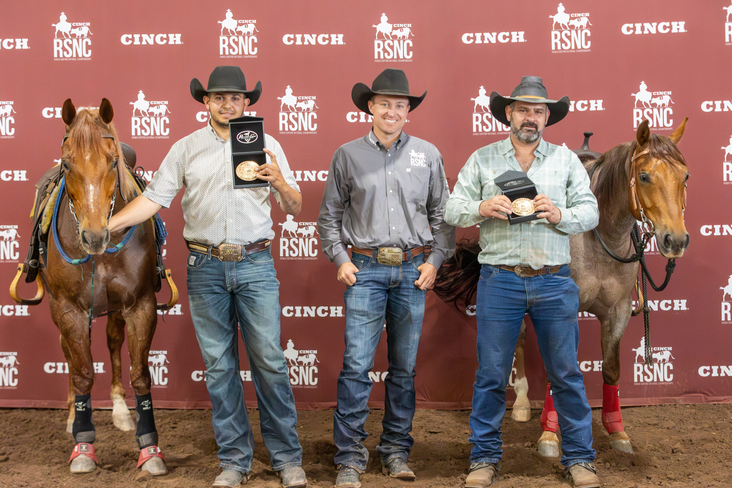 Three cowboys hold championship buckles between two horses in front of a Cinch RSNC backdrop