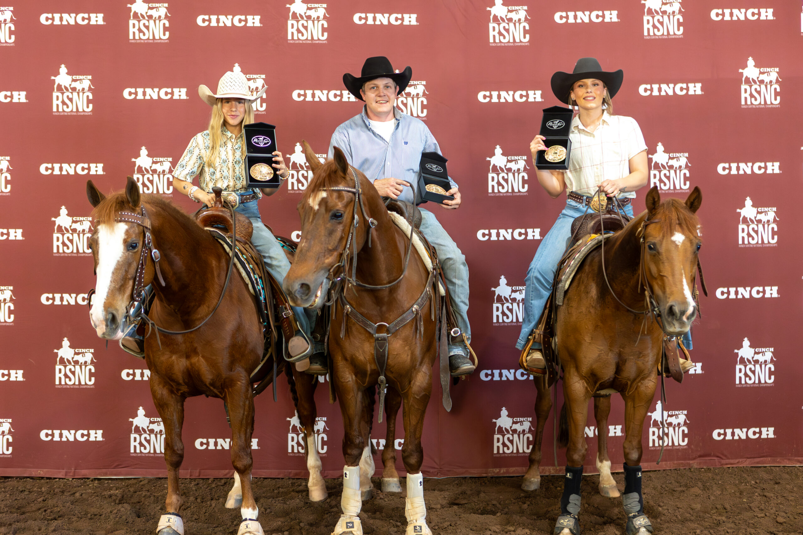 Three riders on sorrel horses hold up championship buckles in presentation boxes before a Cinch RSNC backdrop