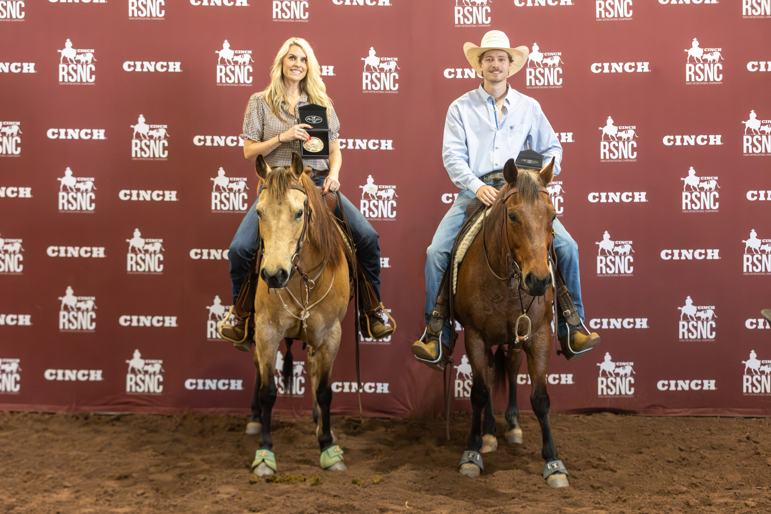 Two riders hold champion buckle cases on horseback in front of a maroon Cinch RSNC backdrop in a sandy arena