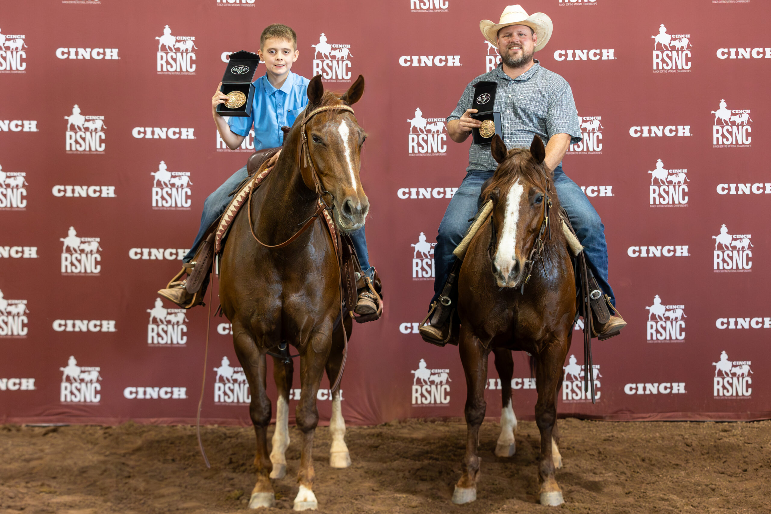 A young rider and a man in a cowboy hat hold up championship buckles on horseback before a Cinch RSNC backdrop