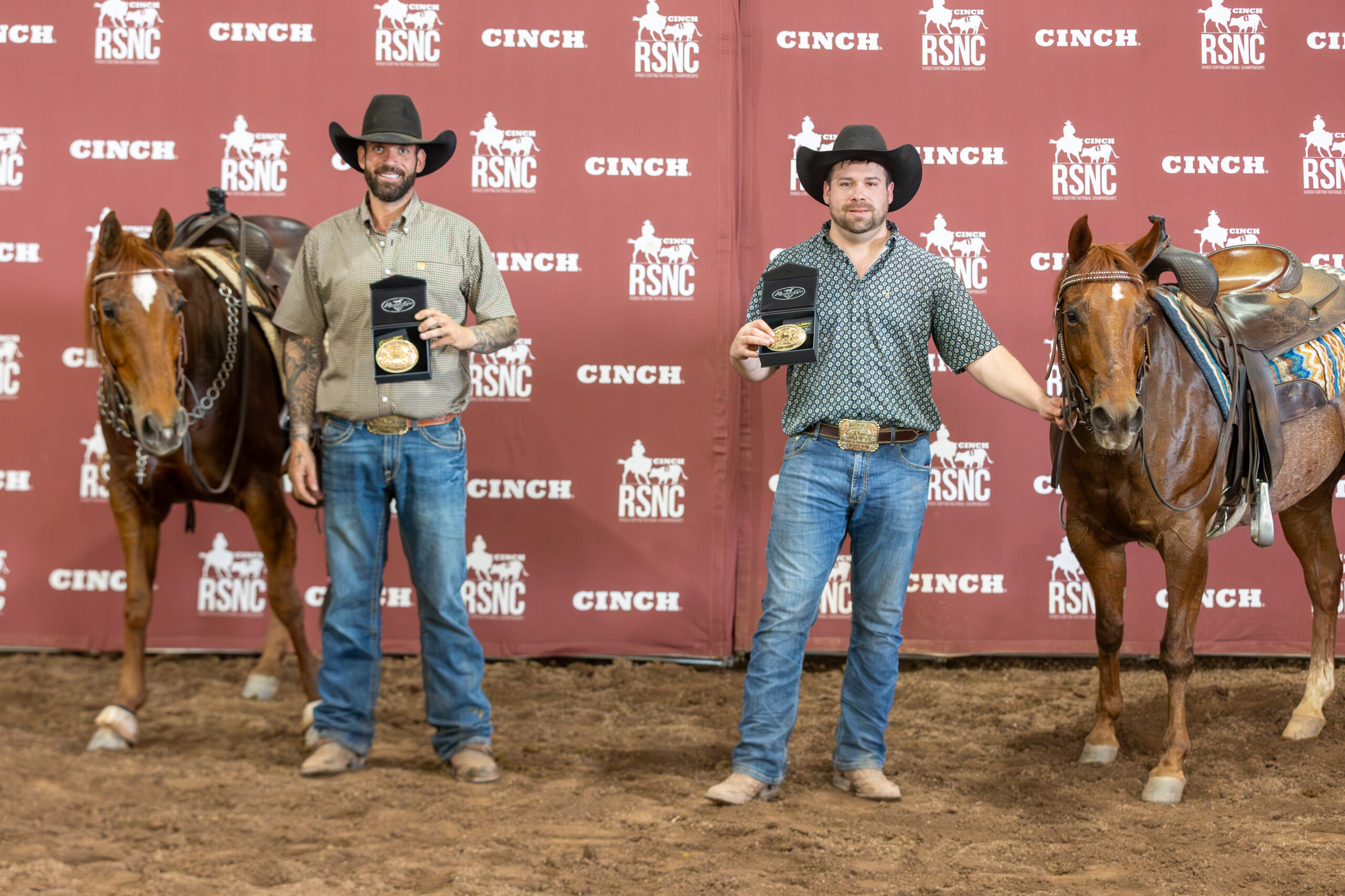 Two cowboys in black hats hold up champion belt buckles in presentation cases beside their saddled sorrel horses in front of a Cinch RSNC backdrop