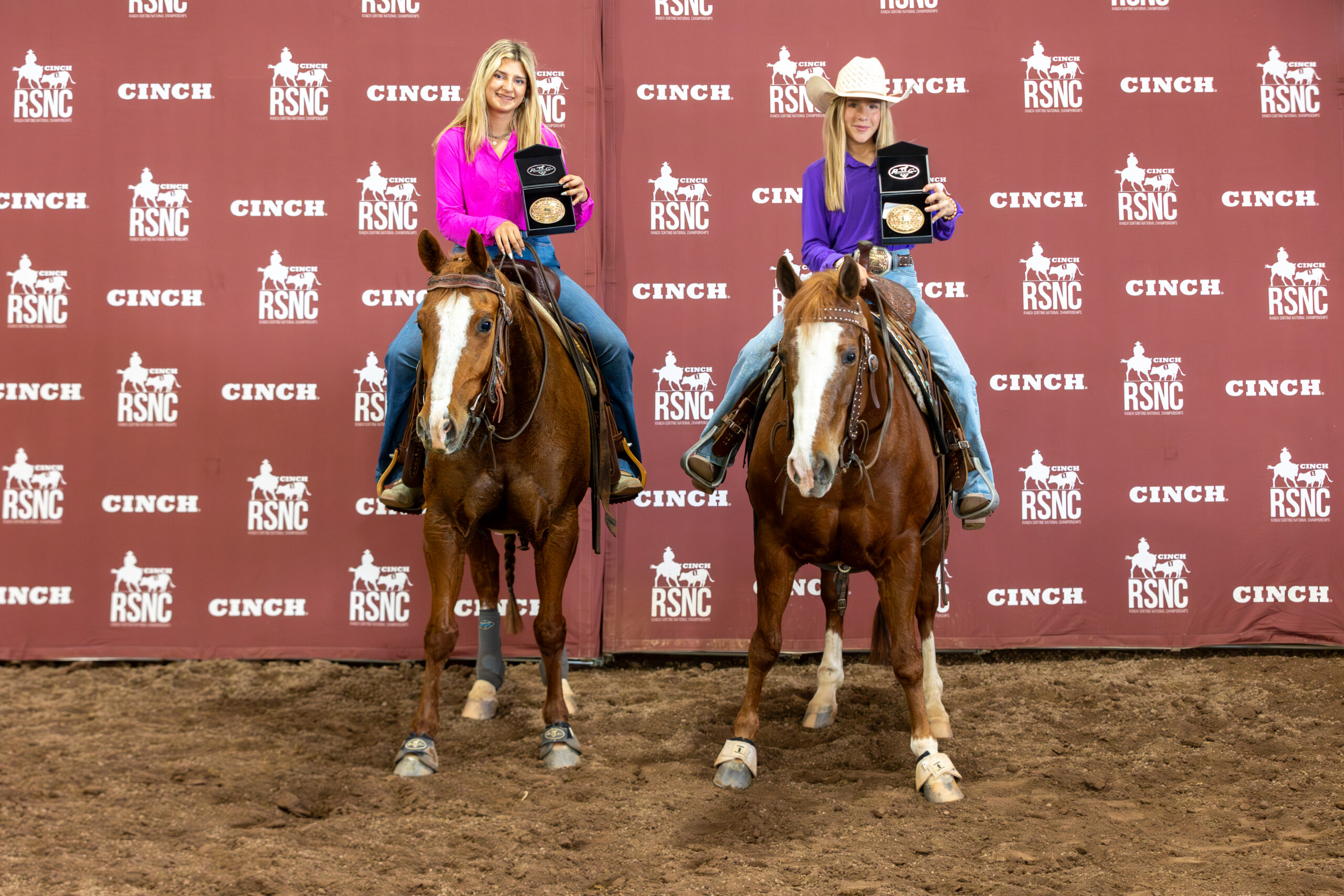 Two young riders on sorrel horses hold up championship buckles in presentation boxes in front of a Cinch RSNC backdrop
