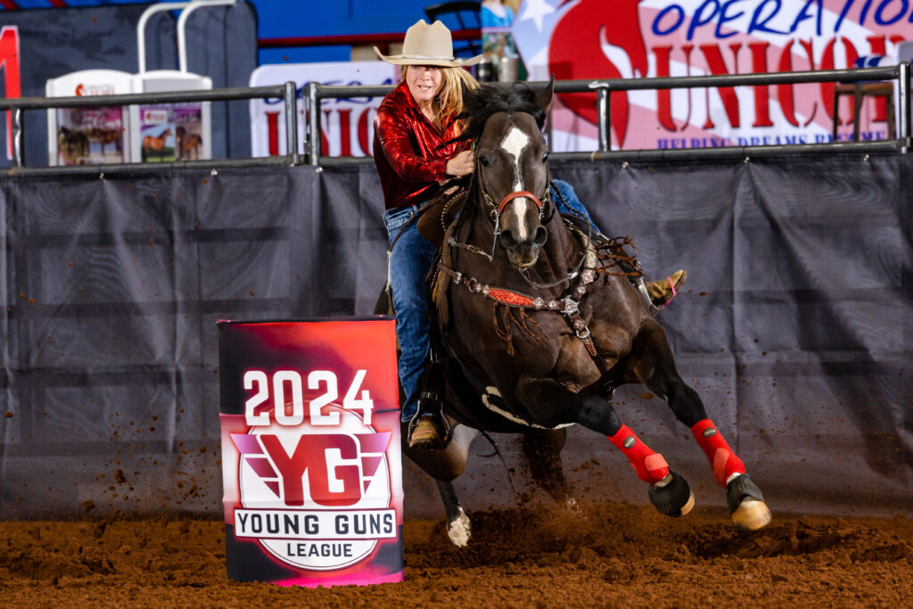 Barrel racer in a red shirt and cowboy hat rounds a 2024 Young Guns League barrel on a dark horse in red splint boots