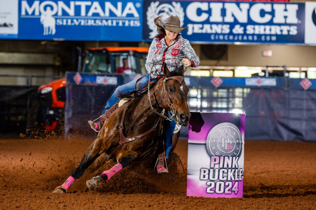 Barrel racer in a cowboy hat rounds a Pink Buckle 2024 barrel on a bay horse in pink polo wraps, dirt flying