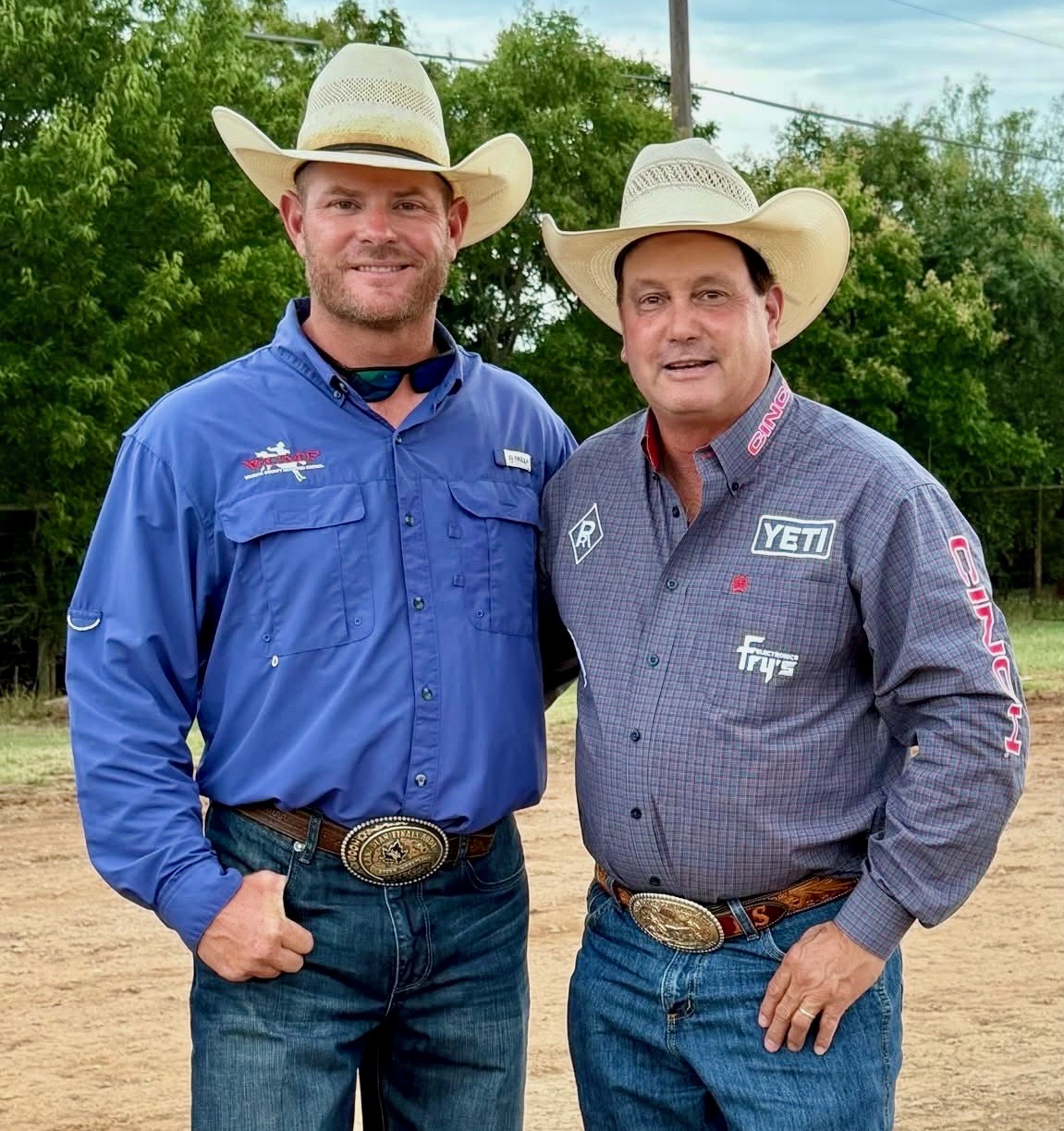 Two men in cowboy hats and western wear with sponsor logos stand together at a rodeo arena